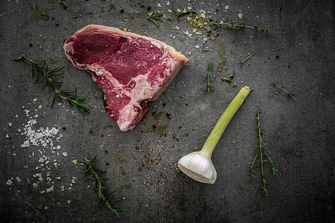 Raw t-bone steak, rosemary, thyme, halved garlic bulb, spices and sea salt