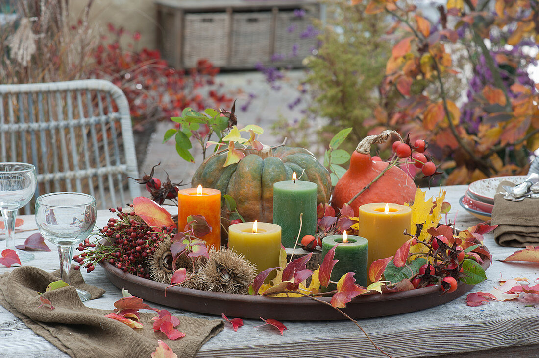 Tischdekoration mit Kerzen, Kürbis, Hagebutten, Maronen und Herbstlaub auf großem Untersetzer