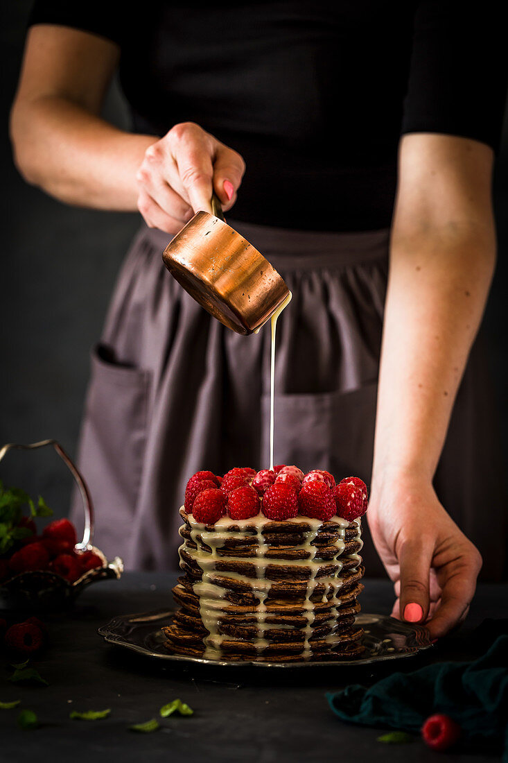 A woman pouring vanilla sauce onto a stack of chocolate pancakes topped with fresh raspberries