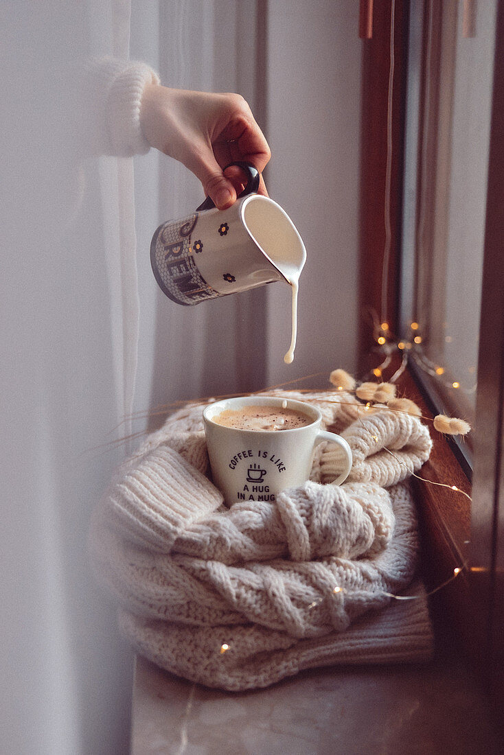 Frau gießt Milch in eine Tasse Kaffee (Winterstimmung)