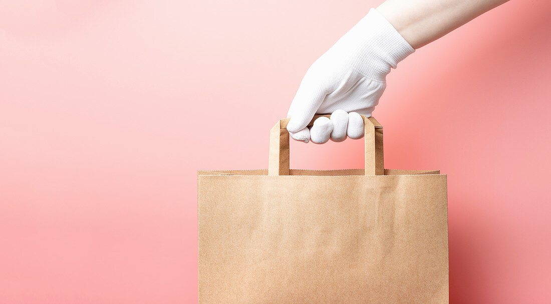 Female hand in a white glove holds brown cardboard bag on a pink background - food delivery concept