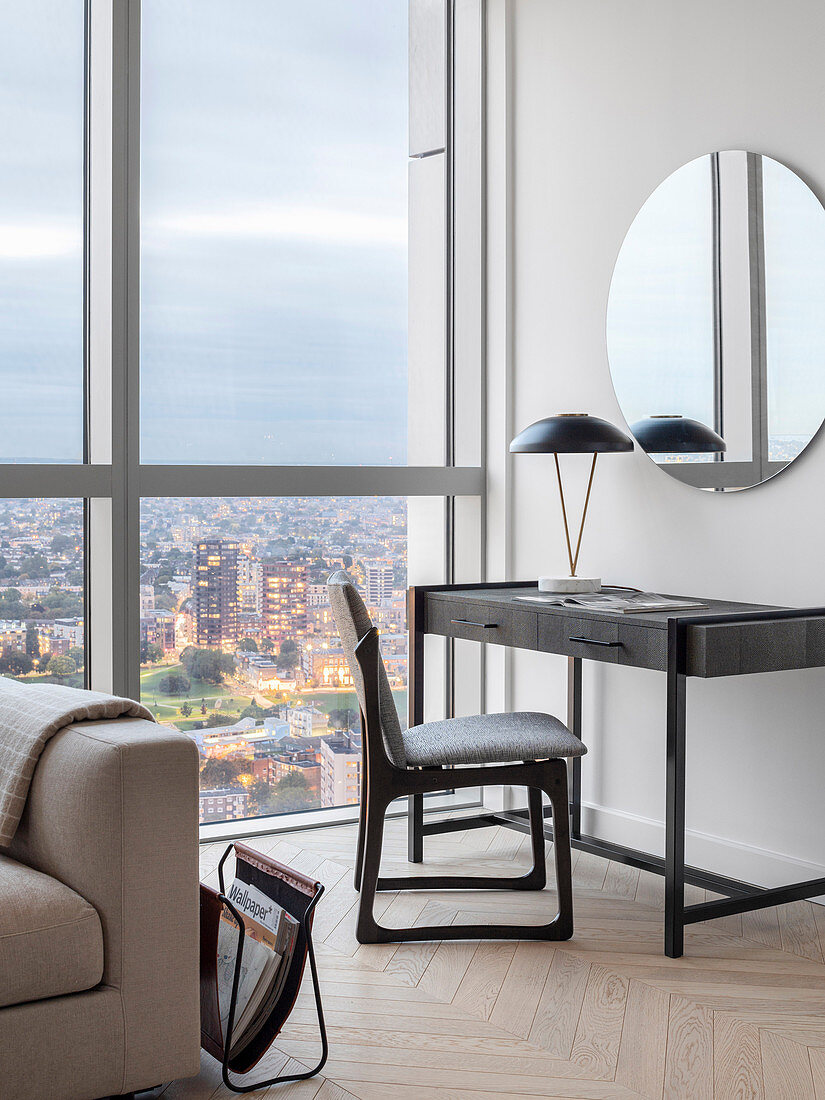 Desk and round mirror next a floor-to-ceiling window in a high-rise building