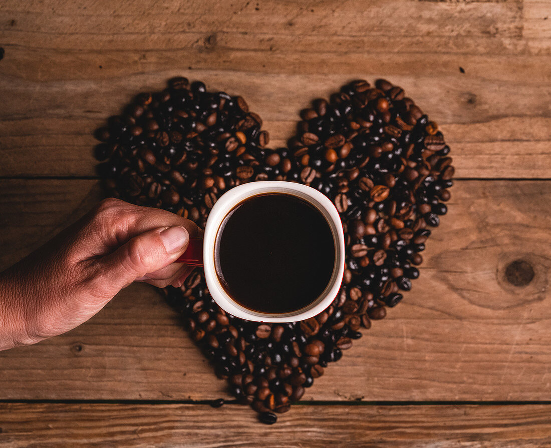 Hand mit einer Tasse Kaffee und Kaffeebohnen in Herzform arrangiert