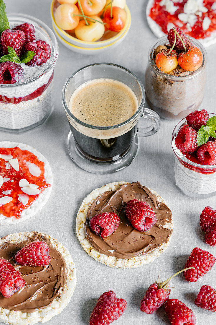 Glass cup of coffee with chia pudding garnished with raspberries and white cherries with wheat crispbreads with nut spread and jam with almond pieces