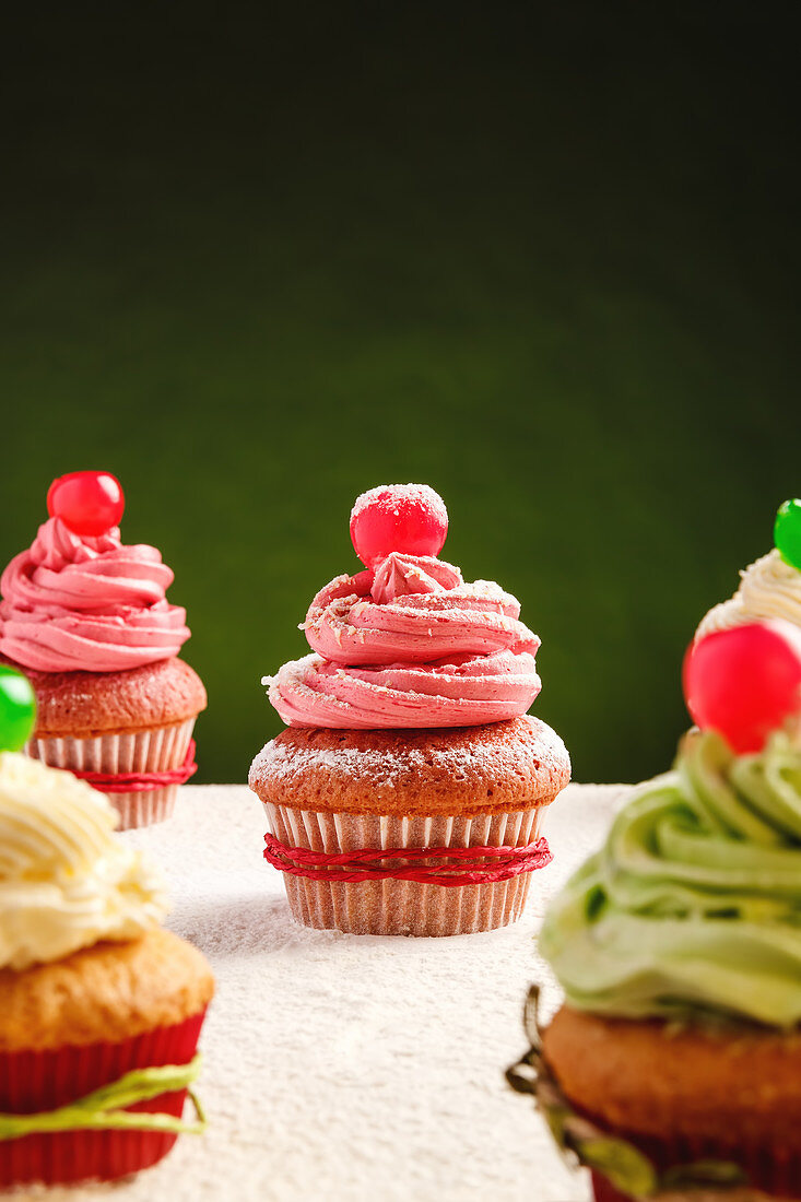 Yummy festive Christmas cupcakes decorated with colorful frosting and cherry berries placed on table sprinkled with white sugar powder against green background