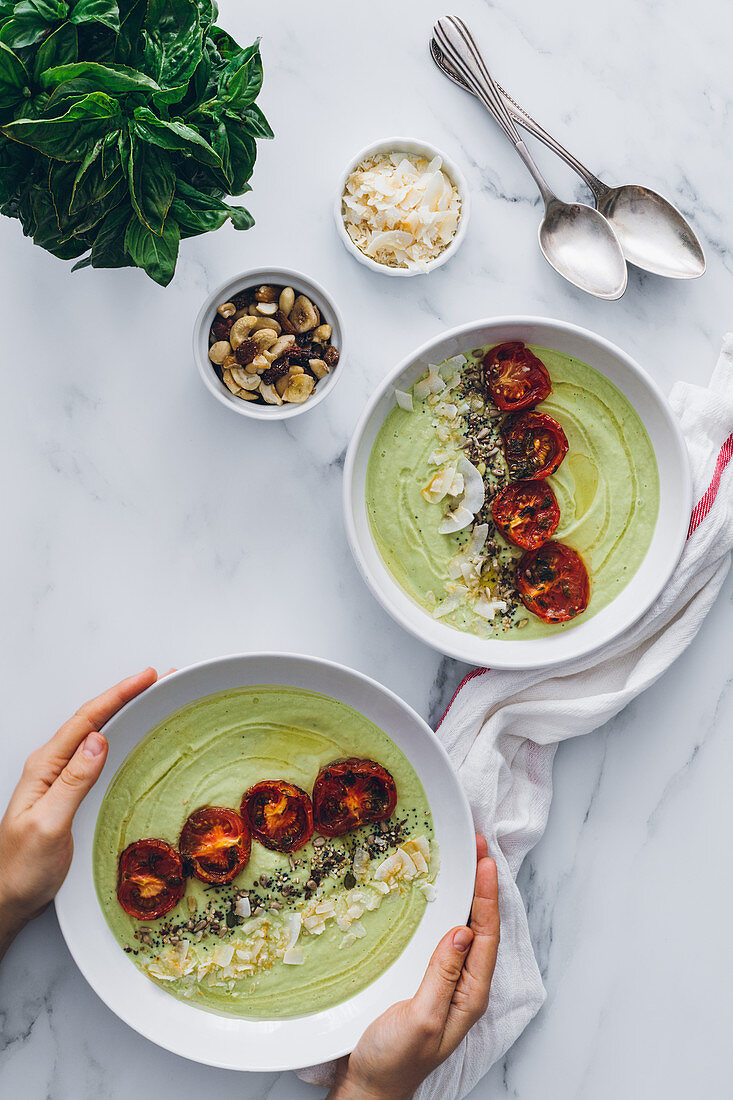 Crop unrecognizable person serving bowls with delicious avocado and apple cream soup decorated with sun dried tomatoes and toasted grated coconut with cashew nuts and fresh basil