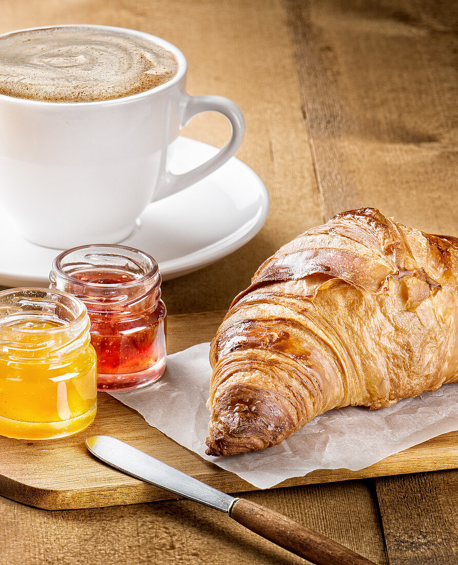 Freshly baked croissant served with fruit marmalade and cup of hot coffee with milk on wooden table