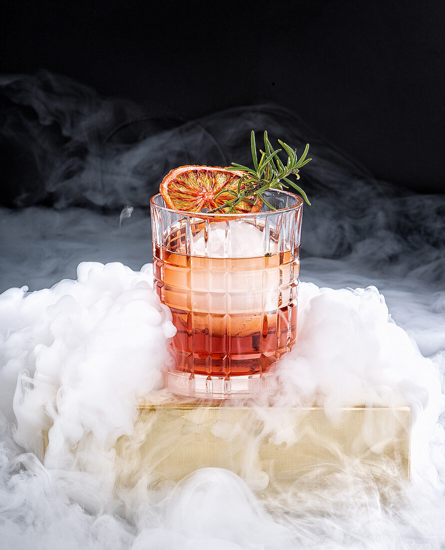 Glass with refreshing alcohol cocktail with red orange and ice garnished with fresh rosemary