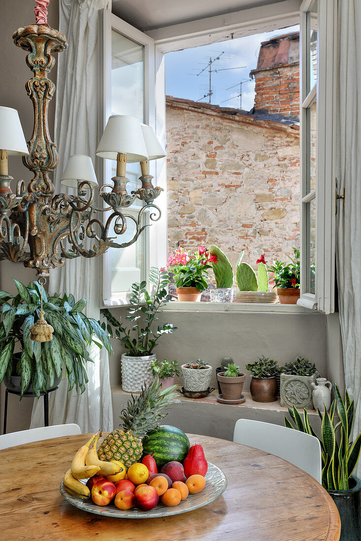 Ornate chandelier above plate of fruit on round antique table and potted plants in background