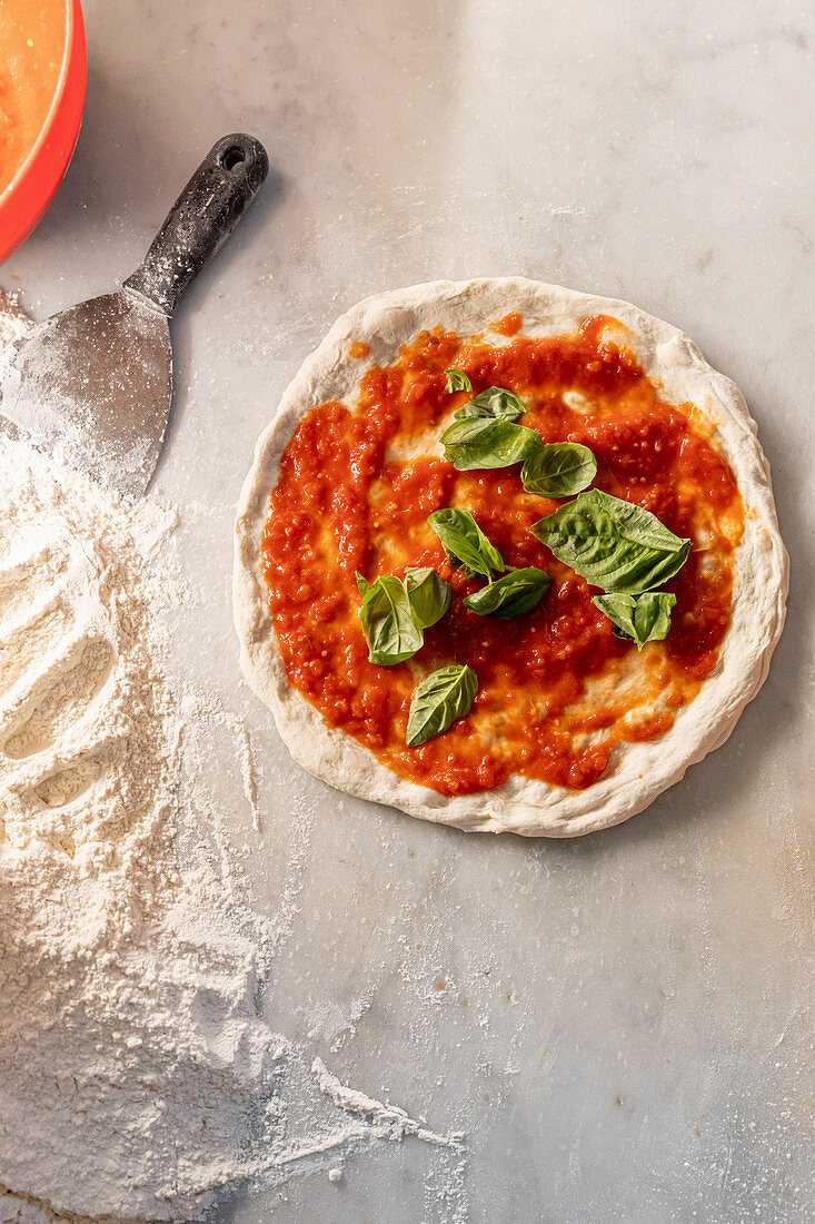 A pizza being made with tomato sauce and basil leaves