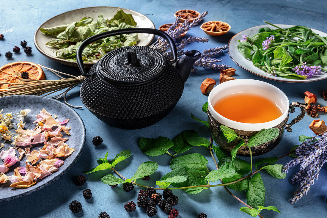 Cup of herbal tea with a tea pot and an assortment of ingredients, herbs, fruits and flowers