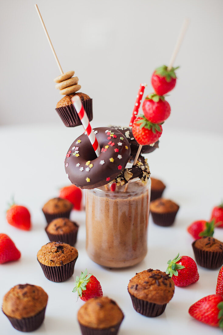 Glass of chocolate milkshake garnished with fresh strawberries and assorted sweet pastry on white table