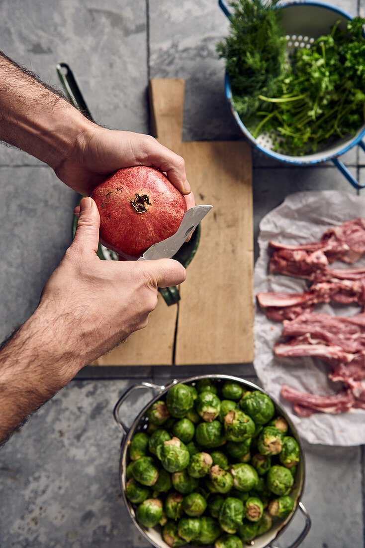 A pomegranate being sliced next to Brussels sprouts, lamb chops and herbs