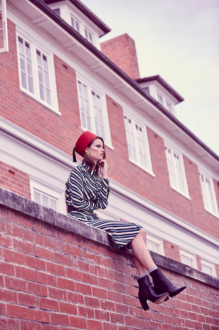 A young woman wearing a 1960s outfit with a hat sitting on a brick wall