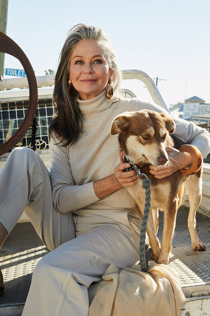 A grey-haired woman wearing a turtleneck jumper and light trouser sitting on a pick-up truck with a dog