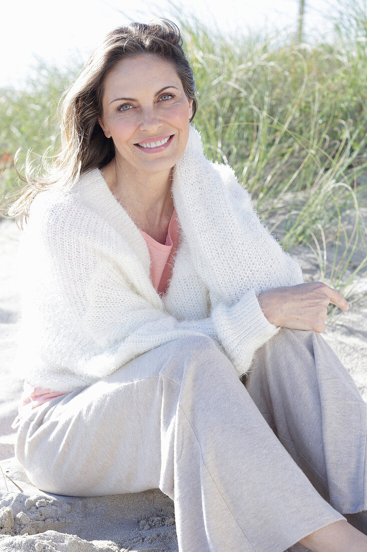 A long-haired woman sitting in the sand on the beach wearing a light jumper and trousers