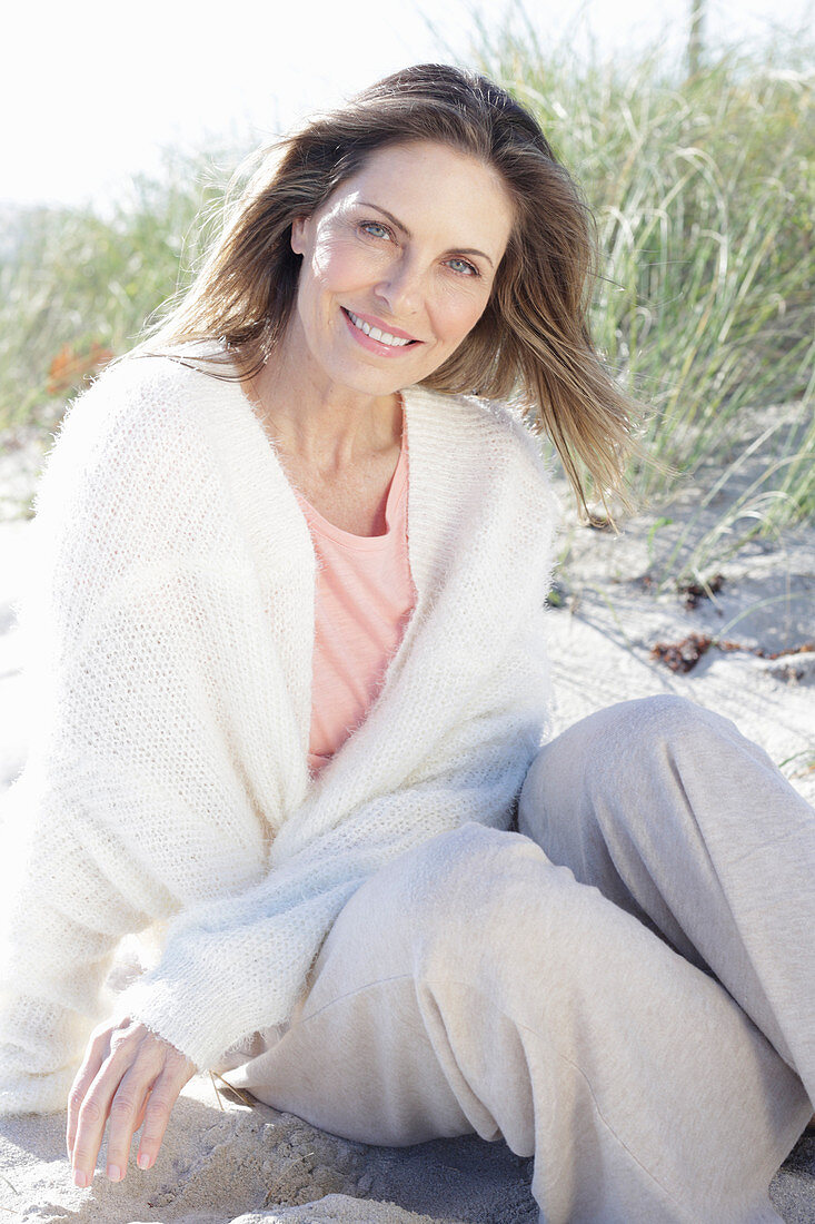 A long-haired woman sitting in the sand on the beach wearing a light jumper and trousers