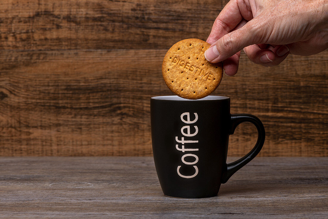 Person holding cookie over black mug with hot coffee