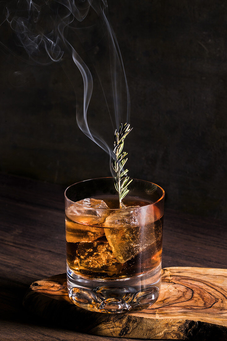 Old fashioned cocktail with ice cubes and fuming rosemary branch