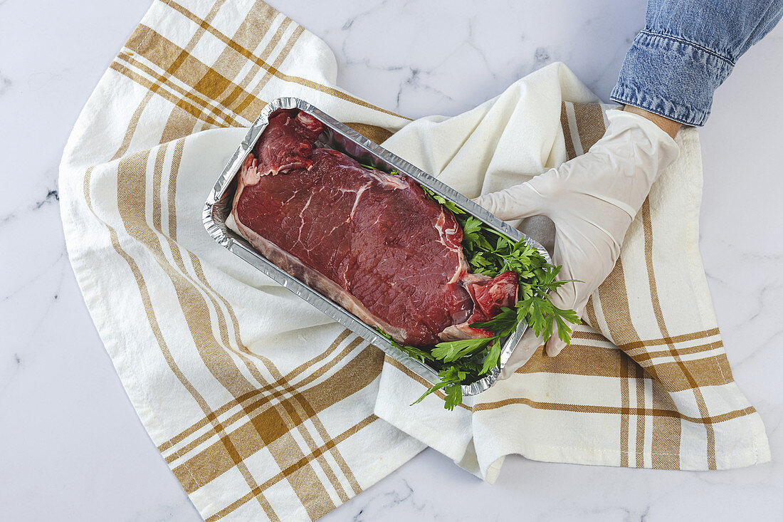 Hand with gloves holds takeaway container with raw steak