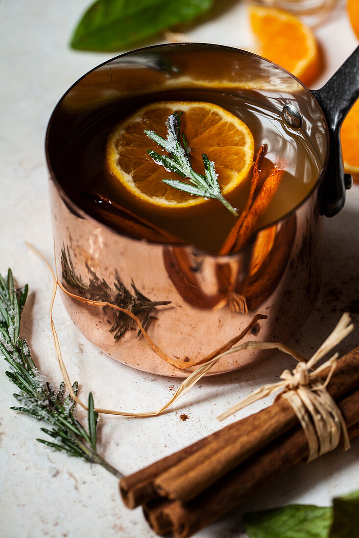 Apple cider heated in an antique copper pot, with oranges with leaves, orange slices, cinnamon sticks and sugared rosemary