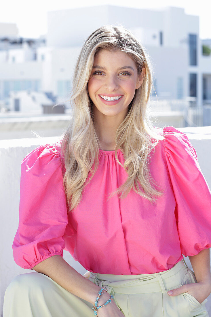 A young blonde woman wearing a pink top with puffed sleeves and white summer trousers