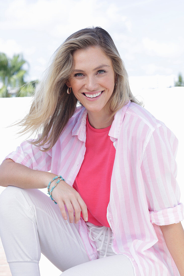 A young blonde woman wearing a pink top, a striped shirt and white trousers