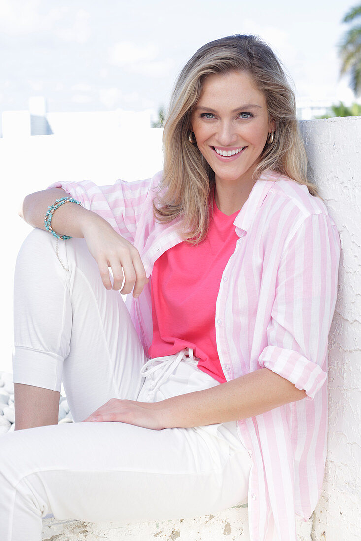 A young blonde woman wearing a pink top, a striped shirt and white trousers