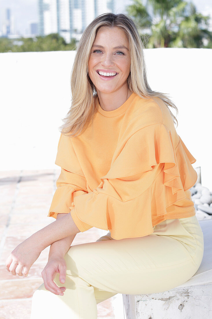 A young blonde woman wearing an apricot blouse and a pair of light trousers