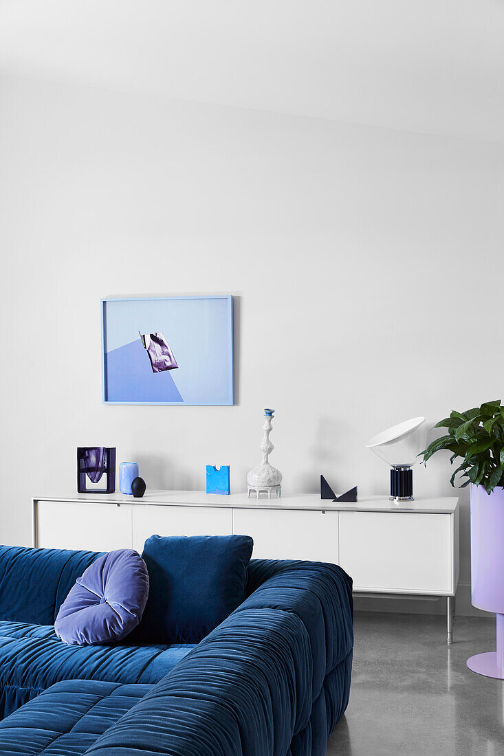Petrolblaues Samtsofa vor weißem Lowboard, darüber moderne Kunst im Wohnzimmer
