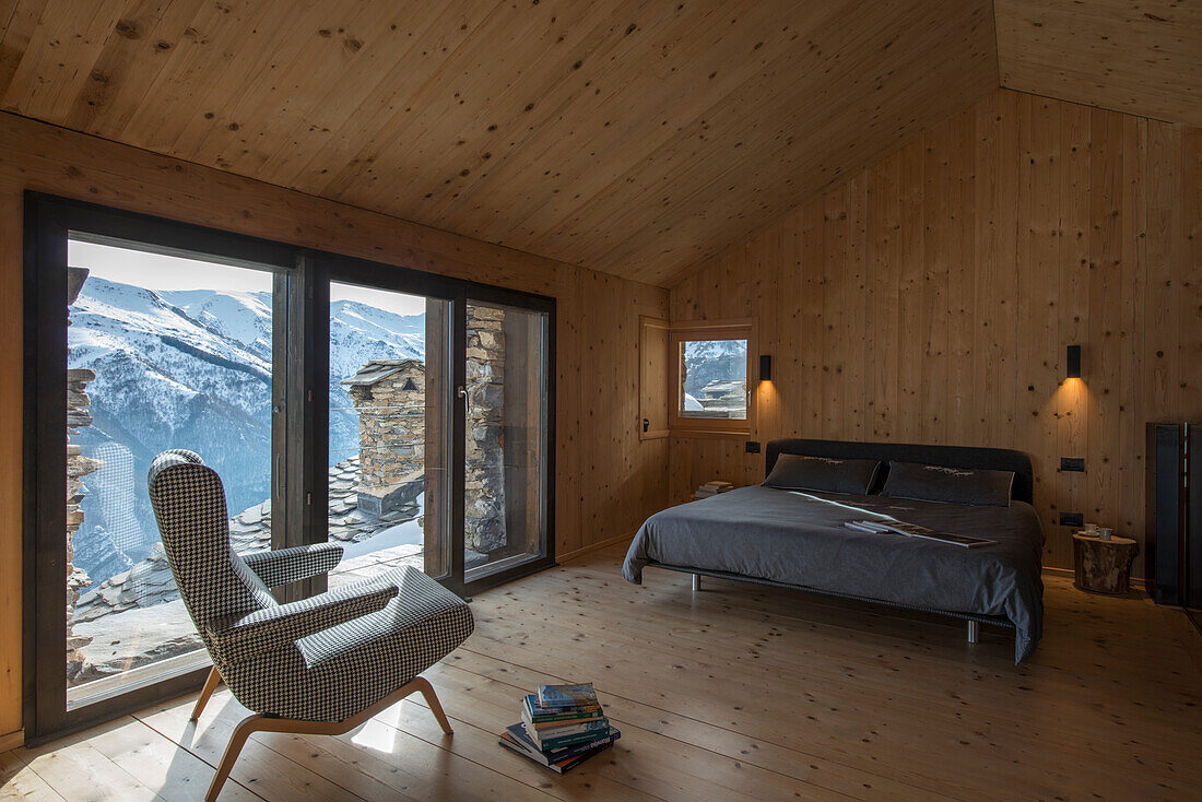 Armchair and double bed next to glass wall in bedroom with wood panelling