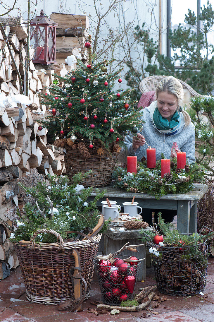 Christmas terrace with Christmas tree, Advent wreath, baskets with fir branches, pinecones and Christmas tree decorations, a mug with hot mulled cider on wooden disc, woman lighting candles, antlers, and old wooden skates as decoration