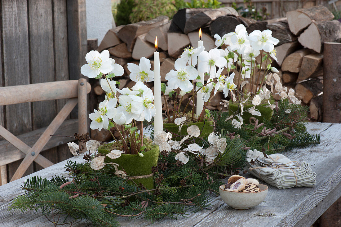 Christmas table decorations on the terrace: candles, Christmas roses in a felt pot, fir branches and silver coins, stacks of firewood in the background