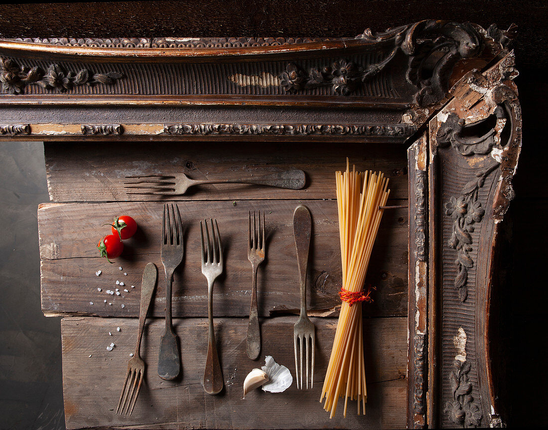 An arrangement of antique forks, spaghetti, garlic and tomatoes in a wooden frame
