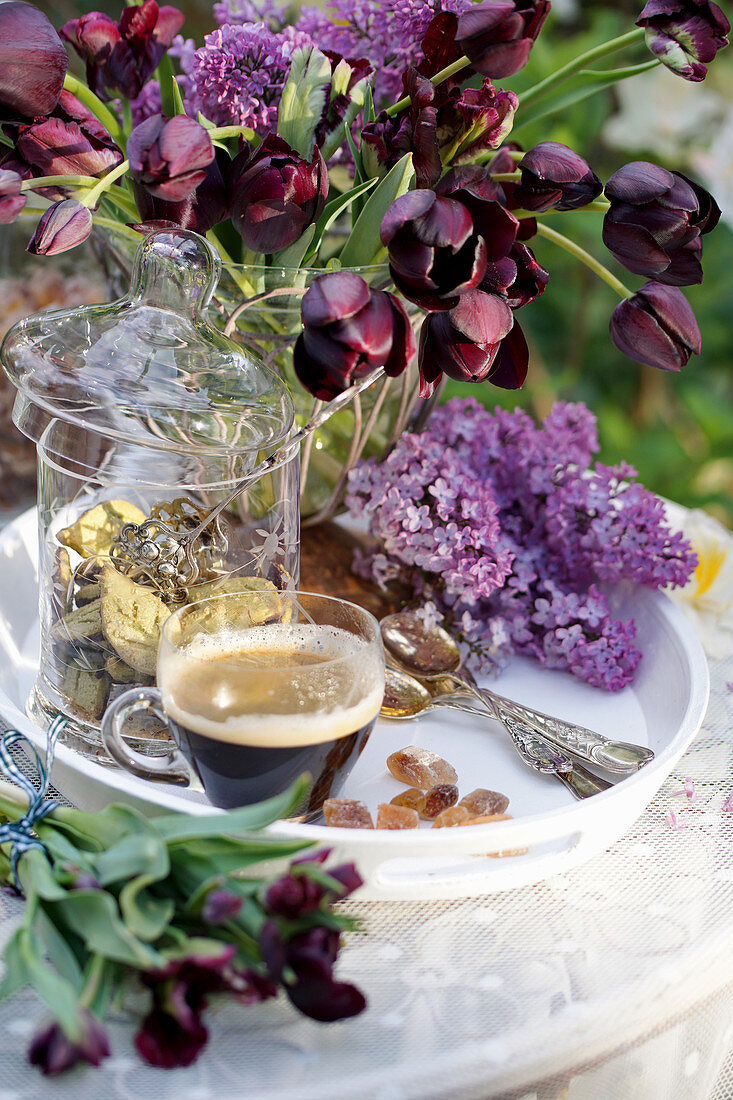 Vase of black tulips, lilac flowers and cup of espresso on tray