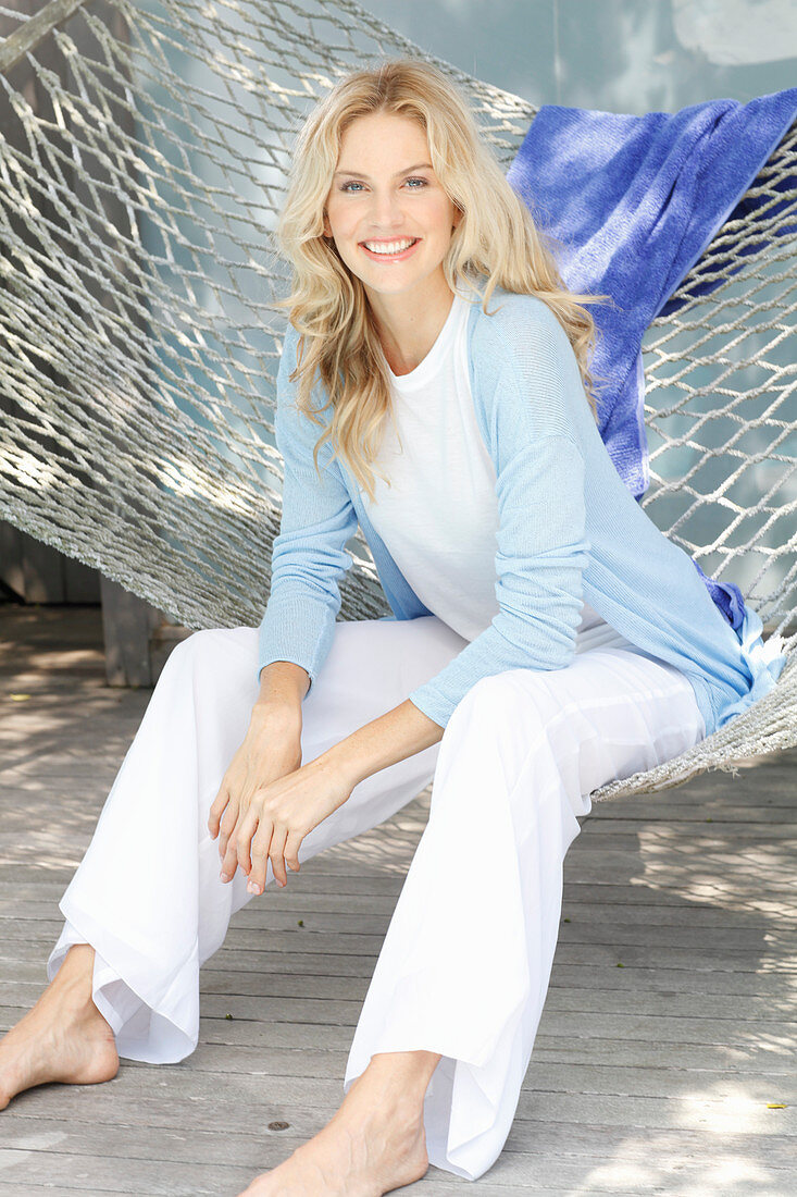 Blonde Frau in weißem Top, weißer Hose und hellblauer Strickjacke sitzt in der Hängematte