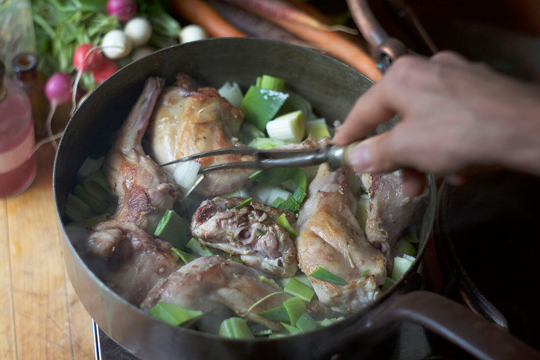 Hand hält Zinkengabel in Kupferpfanne mit gebratenem Kaninchen und Lauch