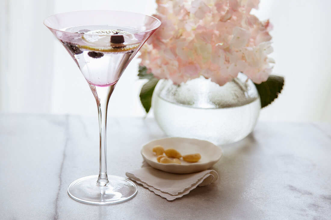 Martini im rosa Glas, Cashewnüsse und Blumenvase auf Marmortisch