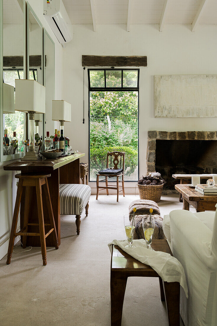 Console with bar stool in front of wall mirror in rustic living room, fireplace and window in background