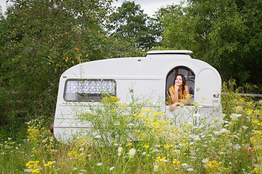 Young woman looking out of caravan in idyllic meadow