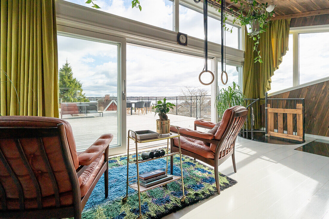 Armchairs with leather upholstery below gymnastic rings in living room with glass wall and terrace view