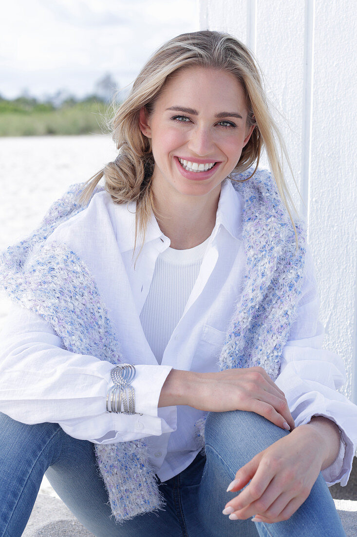 A long-haired blonde woman wearing a white shirt blouse and jeans with a jumper over her shoulders