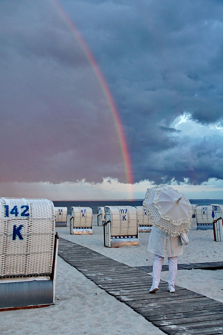 Mädchen mit Regenschirm am Strand mit Strandkörben, Regenbogen im Hintergrund