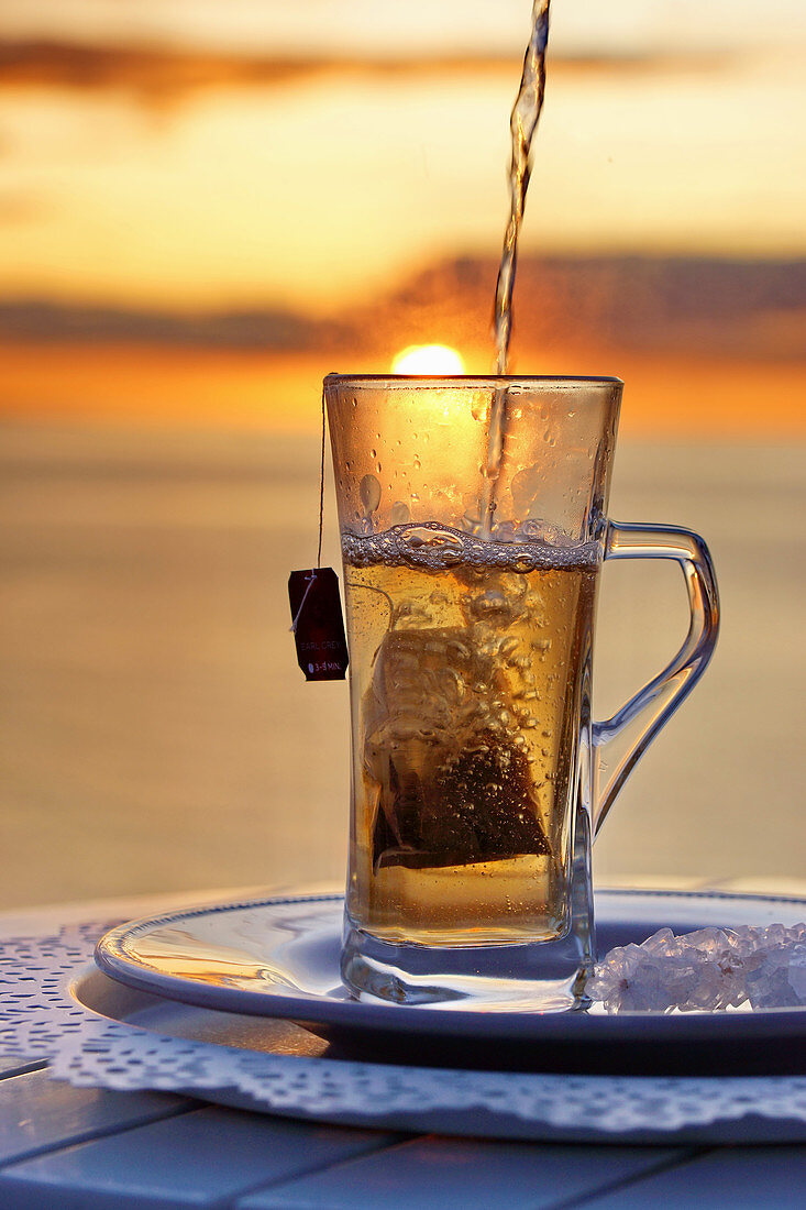 Heißer Tee mit Teebeutel im Glas, am Meer vor Sonnenuntergang