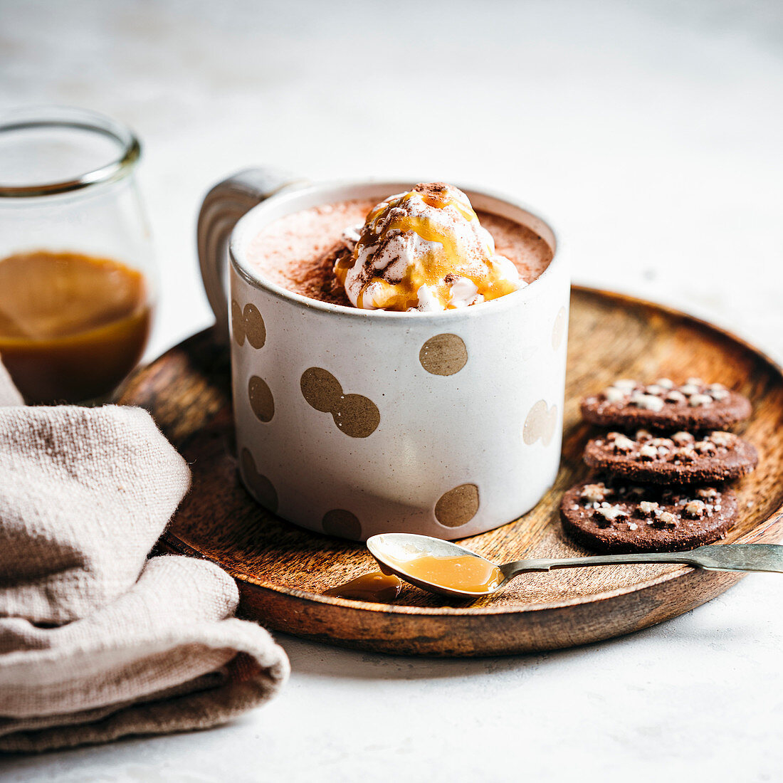 Mug of hot chocolate, topped with caramel drizzled whipped creme, served on a plate with chocolate cookies