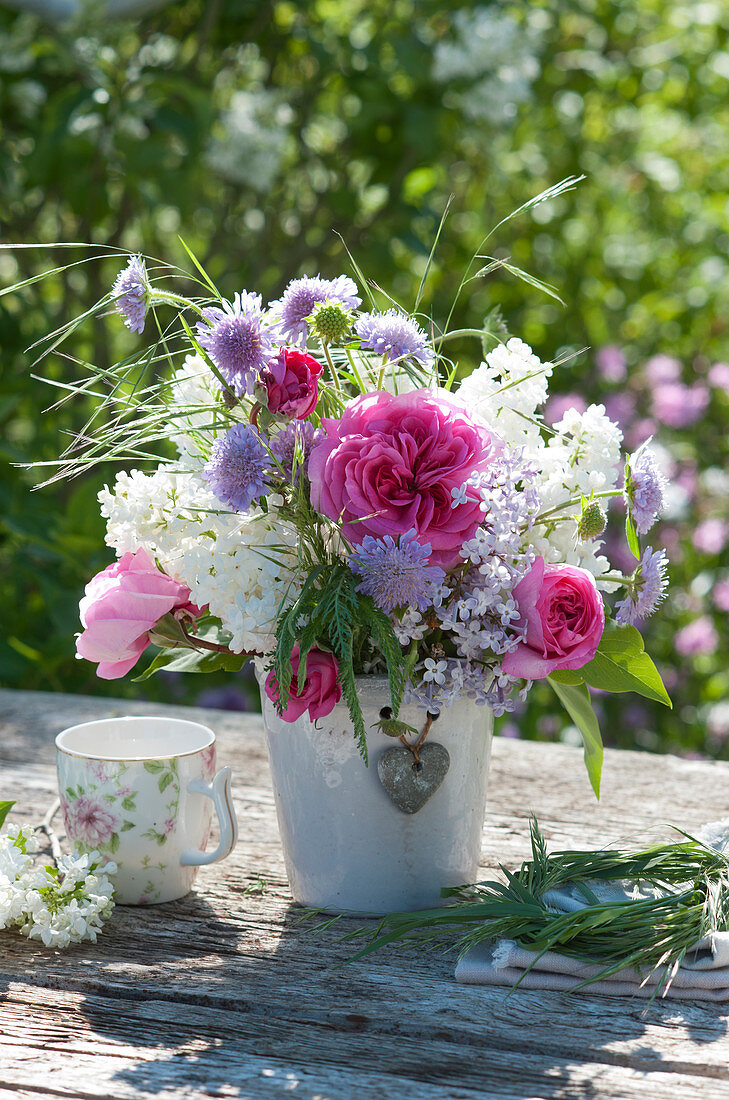 Fragrant bouquet of lilacs, rose blossoms, and Knautia with grass and yarrow leaves in a flower vase with heart pendant