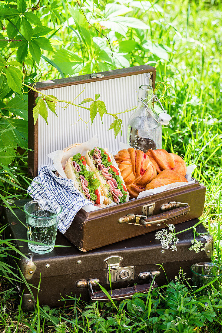 Vintage Picknickkoffer mit Basilikumpesto, Schinken-Sandwiches, Gebäck und Sprudelwasser