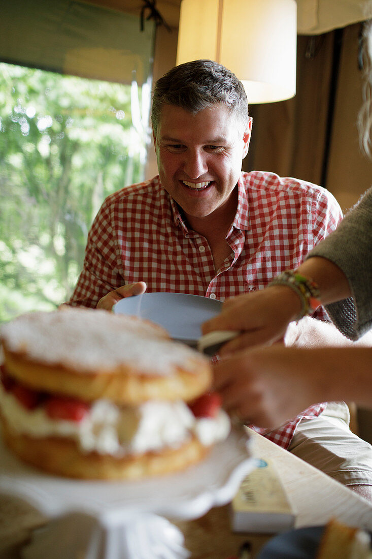 Happy man watching wife slice and serve strawberry cake