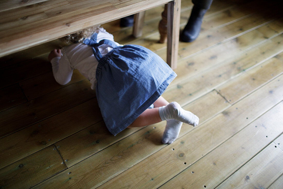 Girl in denim dress crawling under wood dining bench