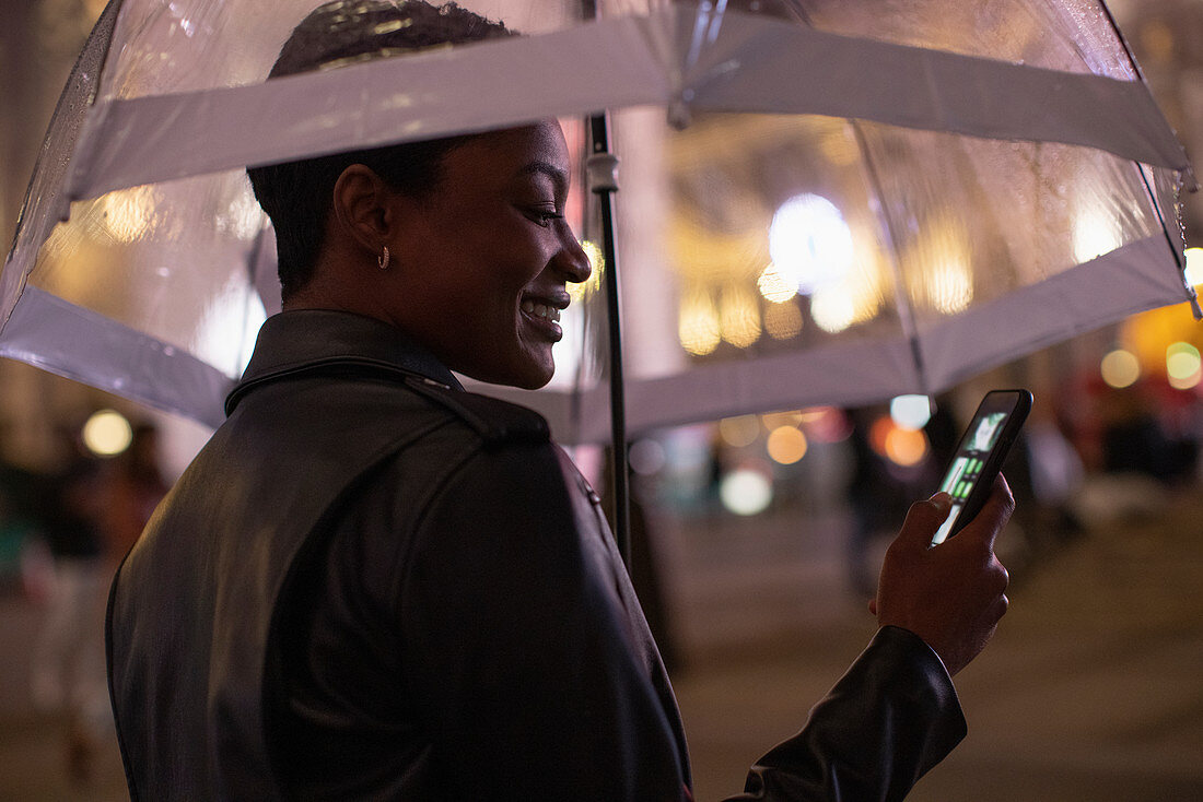 Happy young woman using smartphone under umbrella at night